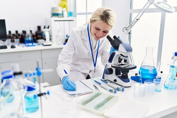 Νεαρή Ξανθιά Γυναίκα Επιστήμονας Χρησιμοποιώντας Μικροσκόπιο Κρατώντας Σημειώσεις Στο Εργαστήριο — Φωτογραφία Αρχείου