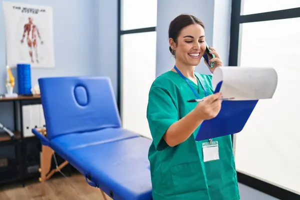 Young Beautiful Hispanic Woman Physiotherapist Reading Document Talking Smartphone Rehab — стоковое фото