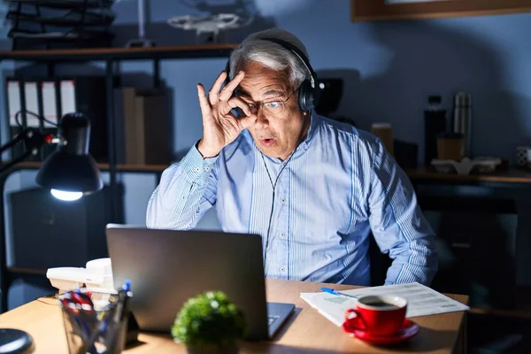 Hispanic senior man wearing call center agent headset at night doing ok gesture shocked with surprised face, eye looking through fingers. unbelieving expression.