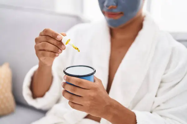 Young latin man relaxed on sofa with facial mask treatment drinking coffee at home