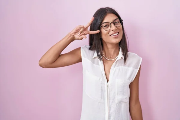 ブルネット若い女性がピンクの背景に立っている眼鏡をかけて顔の上に指で平和のシンボルをやって 笑顔明るい勝利を示す — ストック写真