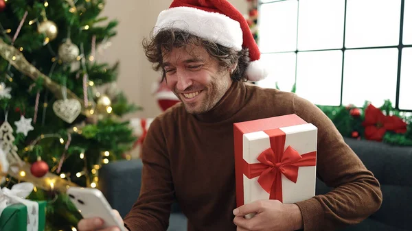 自宅でクリスマスプレゼントを持ってソファーに座ってスマートフォンを使用する若いヒスパニック男性 — ストック写真