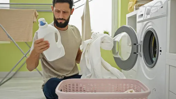 Young hispanic man washing clothes holding towel and detergent bottle at laundry room