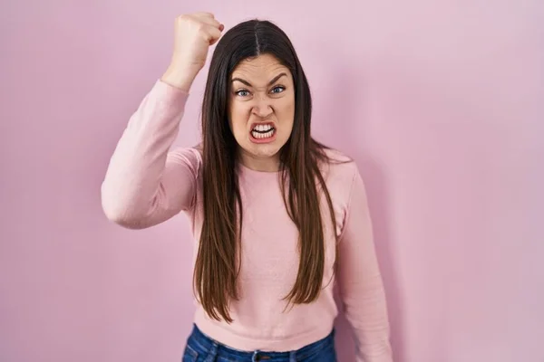ピンクの背景に立っている若いブルネットの女性は怒りで叫んでいる間に怒りと怒りに苛まれ 激怒して拳を上げる 怒りと攻撃的な考え方 — ストック写真