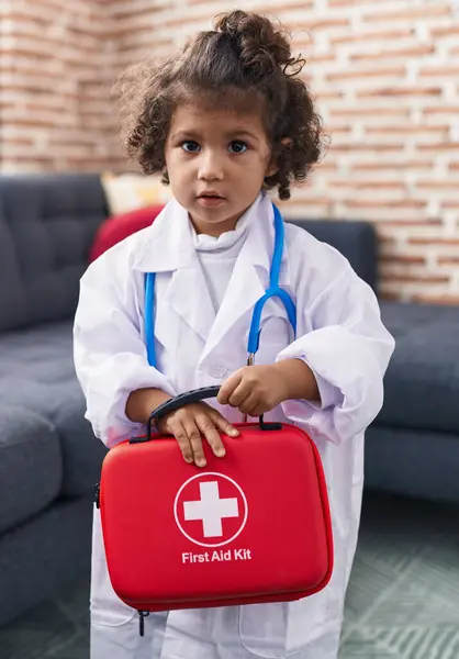 Adorable hispanic girl wearing doctor uniform holding first aid kit box at home