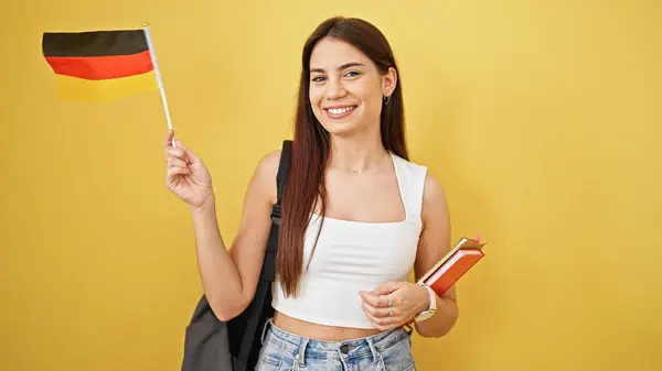 若い美しいヒスパニックの女性学生は 孤立した黄色の背景の上に本やドイツの旗を保持する自信に微笑みます — ストック写真