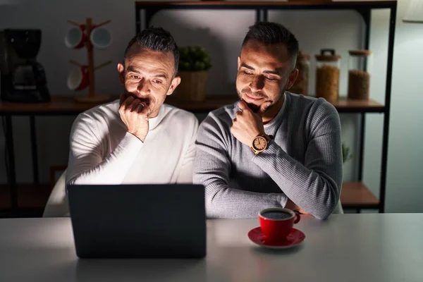 Homosexual couple using computer laptop looking confident at the camera smiling with crossed arms and hand raised on chin. thinking positive.