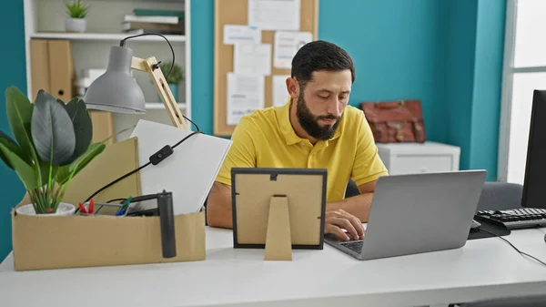 Young hispanic man being fired sitting on table using laptop at the office