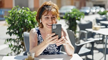 Orta yaşlı bir kadın kahve dükkanının terasında gülümseyen bir video mesajı alıyor.