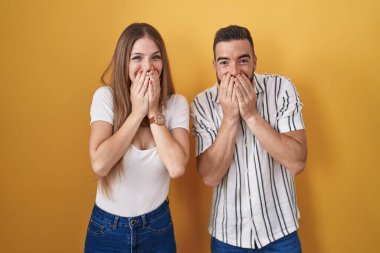 Sarı arka planda dikilen genç çift gülüyor ve utanıyor kıkırdıyor elleri, dedikoduları ve skandalları ağızlarıyla örtüyor. 