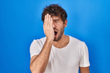 Hispanic young man standing over blue background yawning tired covering half face, eye and mouth with hand. face hurts in pain. 