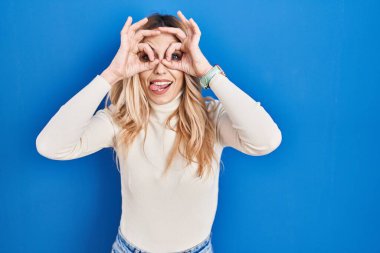 Mavi arka planda dikilen genç beyaz kadın dürbün gibi jest yapıyor. Dürbün gibi dilini çıkartıyor, gözler parmaklarından bakıyor. çılgın ifade. 