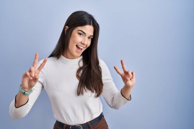 Mavi arka planda dikilen esmer kadın iki elini de zafer işareti yaparak parmaklarını göstererek gülümsüyor. İki numara.. 