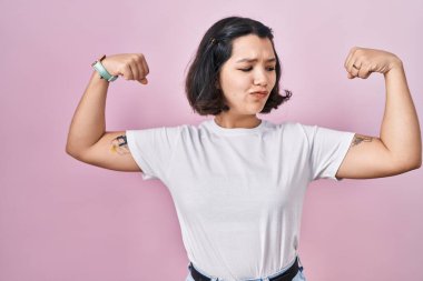 Pembe arka planda günlük beyaz tişört giyen genç İspanyol kadın gururla gülümseyen kol kaslarını gösteriyor. fitness konsepti. 