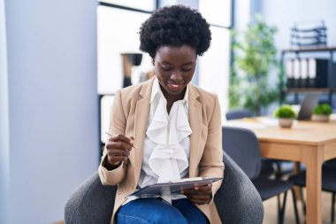 Genç Afro-Amerikan kadın iş adamı. Ofisteki sandalyede oturmuş evrak işleriyle uğraşıyor.
