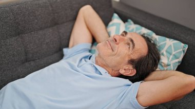 Orta yaşlı bir adam evinde kanepede yatarken elleri başının üstünde rahatlamış.