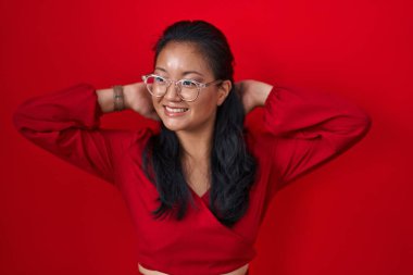 Asyalı genç bir kadın kırmızı arka planda duruyor rahatlıyor ve esniyor, kolları ve elleri başının ve boynunun arkasında mutlu bir şekilde gülümsüyor. 
