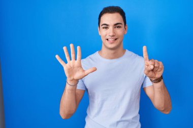 Young hispanic man standing over blue background showing and pointing up with fingers number six while smiling confident and happy. 