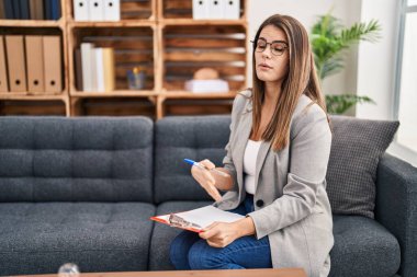 Young beautiful hispanic woman psychologist writing on document at psychology clinic