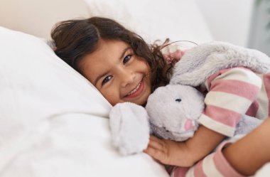 Sevimli İspanyol kız yatak odasında uzanmış oyuncak bebeğe sarılıyor.