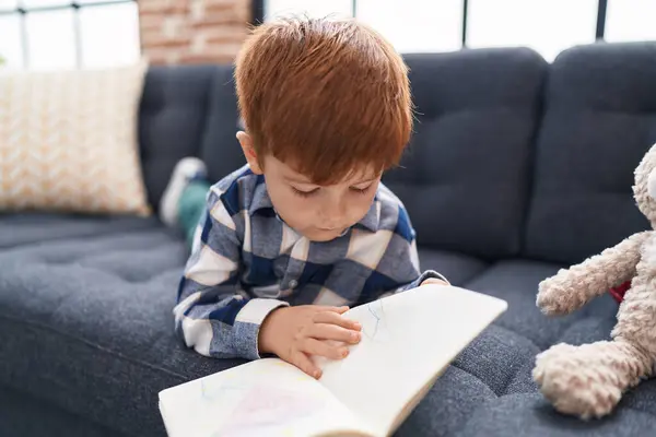 Adorable toddler reading story book lying on sofa at home