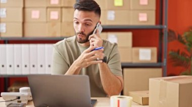 Genç Latin asıllı bir adam, ofisteki bilgisayarını kullanarak akıllı telefondan konuşuyor.