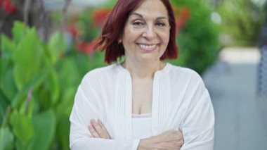 Orta yaşlı bir kadın gülümsüyor. Parkta kollarını kavuşturmuş duruyor.