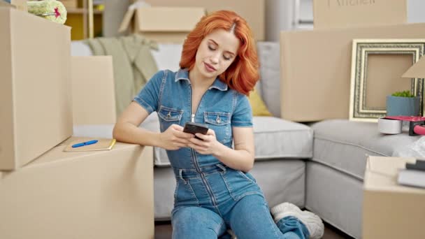 新しい家で床に座っているスマートフォンを使用して若い赤毛の女性 — ストック動画