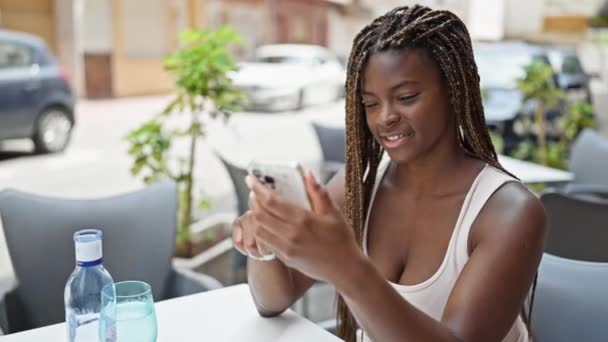 在咖啡店的阳台上 非洲裔美国女人用智能手机坐在桌子上 摆出优胜的姿势 — 图库视频影像