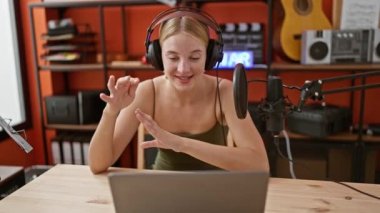 Podcast stüdyosunda kulaklık ve dizüstü bilgisayar kullanarak konuşan genç sarışın kadın radyo muhabiri.