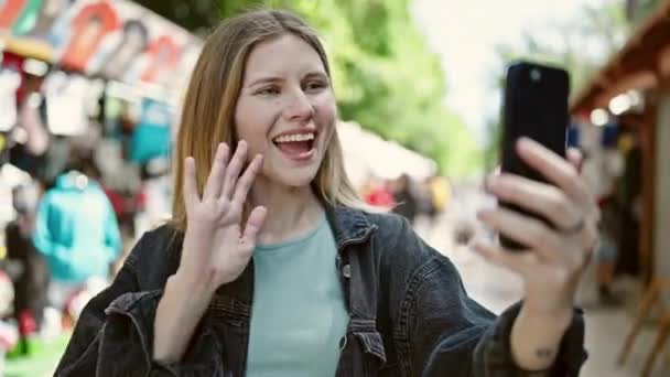 若い金髪の女性は ストリートマーケットでビデオ通話を持っている自信を持って微笑みます — ストック動画