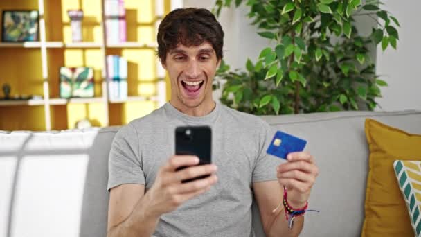 他那拿着智能手机和信用卡在家里沙发上购物的年轻人 — 图库视频影像