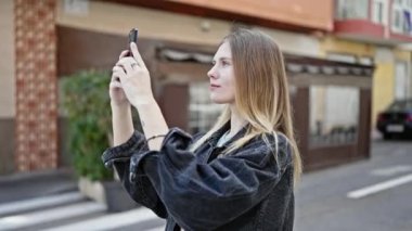 Genç sarışın kadın akıllı telefonun yanında kaset kaydediyor. Kahve dükkanının terasında gülümsüyor.