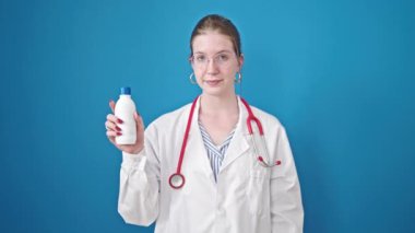 Sarışın kadın doktor gülümsüyor. Kendine güveni tam. Mavi arka planda ilaç şişesi tutuyor.