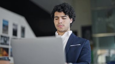 Laptop kullanan genç Latin iş adamı ofise üzgün bakıyor.