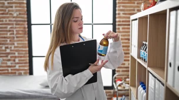 年轻的金发女医生在诊所拿着剪贴板和药瓶 — 图库视频影像
