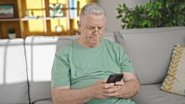 中年白髪男自宅でソファに座ってスマートフォンを使用して — ストック動画