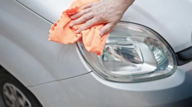 Orta yaşlı, kır saçlı bir adam caddede araba farlarını temizliyor.