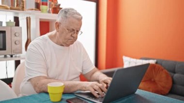 Yemek odasında gülümseyen dizüstü bilgisayar kullanan orta yaşlı gri saçlı bir adam.