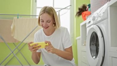 Çamaşır odasında akıllı telefondan video oyunu oynayan genç sarışın kadın.