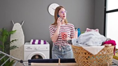 Genç sarışın kadın akıllı telefonda konuşuyor kahve içiyor çamaşır odasında çamaşır makinesini bekliyor.
