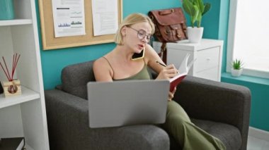 Genç sarışın iş kadını bilgisayarını kullanıyor. Akıllı telefondan tartışıyor. Ofiste not alıyor.