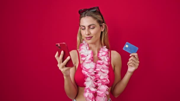 若いですブロンド女性身に着けていますビキニショッピングとともにスマートフォンとクレジットカード上の隔離された赤の背景 — ストック動画