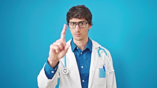 若いヒスパニックの男性医師は 孤立した青い背景の上に指でノーと言う深刻な表現で立っています — ストック動画