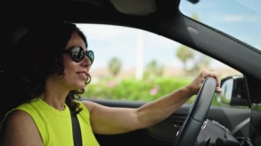 Orta yaşlı İspanyol bir kadın yolda gülümseyen güneş gözlüklü bir araba kullanıyor.