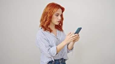 Genç kızıl saçlı kadın, beyaz arka plan üzerinde akıllı telefon kullanarak sürpriz bir yüz ifadesiyle ayakta duruyor.