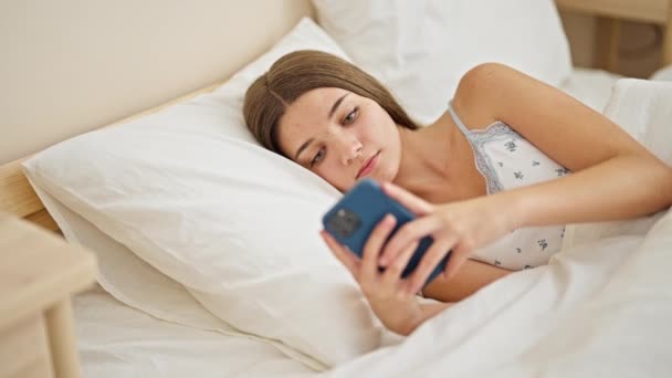 年轻漂亮的女孩用智能手机躺在床上 满脸愁容地看着卧室 — 图库视频影像