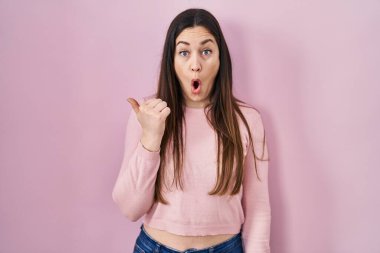 Young brunette woman standing over pink background surprised pointing with hand finger to the side, open mouth amazed expression. 
