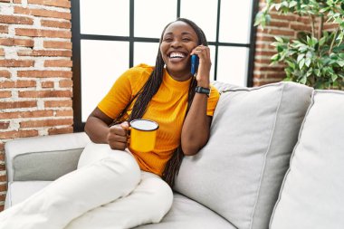 Afro-Amerikalı kadın akıllı telefondan konuşuyor evde kahve içiyor.