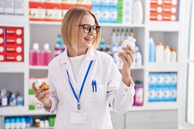 Young blonde woman pharmacist smiling confident holding pills bottles at pharmacy